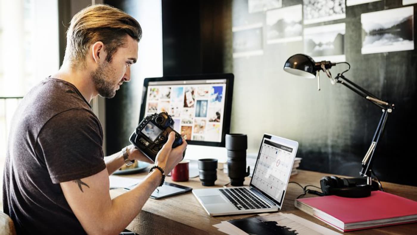 Man working on computer and laptop holding camera